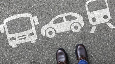 Füße auf Asphalt vor Symbolen für Bus, Auto und Zug als Darstellung verschiedener Verkehrsmitteloptionen.