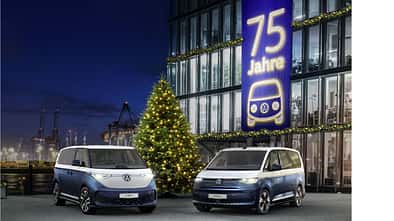 VW ID. Buzz und VW Multivan Sondermodelle vor einem Gebaeude mit Banner „75 Jahre“ und Weihnachtsbaum bei Nacht am Hafen.