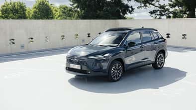 Toyota Corolla Cross in Grau, Front-Dreiviertelansicht, stehendes SUV auf Parkdeck vor Betonwand und Baeumen.