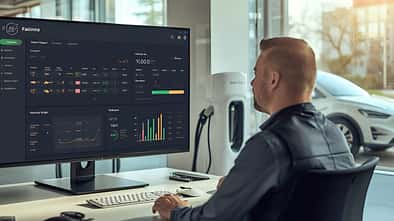 Mann sitzt an einem Arbeitsplatz und analysiert Fuhrparkdaten auf einem Bildschirm, im Hintergrund ein Elektroauto an einer Ladestation