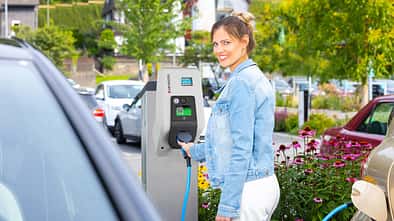 Frau hält Ladekabel an Mennekes AMTRON Wallbox auf einem Parkplatz im Freien neben einem Elektroauto.