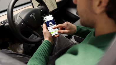 Dienstwagen-Fahrer nutzt Smartphone mit Driver App für E-Auto-Flotte im Elektroauto und sieht Fahrzeugdaten wie Ladezustand und Reichweite.