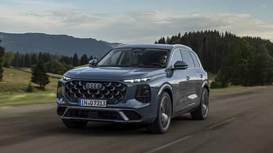Audi Q7 während der Fahrt schräg von vorn auf einer Landstraße mit Wald und Bergen im Hintergrund.