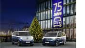 VW ID. Buzz und VW Multivan Sondermodelle vor einem Gebaeude mit Banner „75 Jahre“ und Weihnachtsbaum bei Nacht am Hafen.