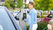Frau hält Ladekabel an Mennekes AMTRON Wallbox auf einem Parkplatz im Freien neben einem Elektroauto.