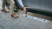 Eine Person lädt ein Elektroauto über einen im Bordstein integrierten Ladepunkt am Fahrbahnrand.