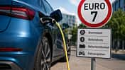 Elektroauto an einer Ladesäule neben einem Euro-7-Schild mit Hinweisen auf Bremsstaub, Reifenabrieb und Fahrzeugdaten.