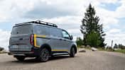 Ford E-Transit Custom AWD Trail in Heckansicht mit Dachträger auf einem Parkplatz in natürlicher Umgebung.